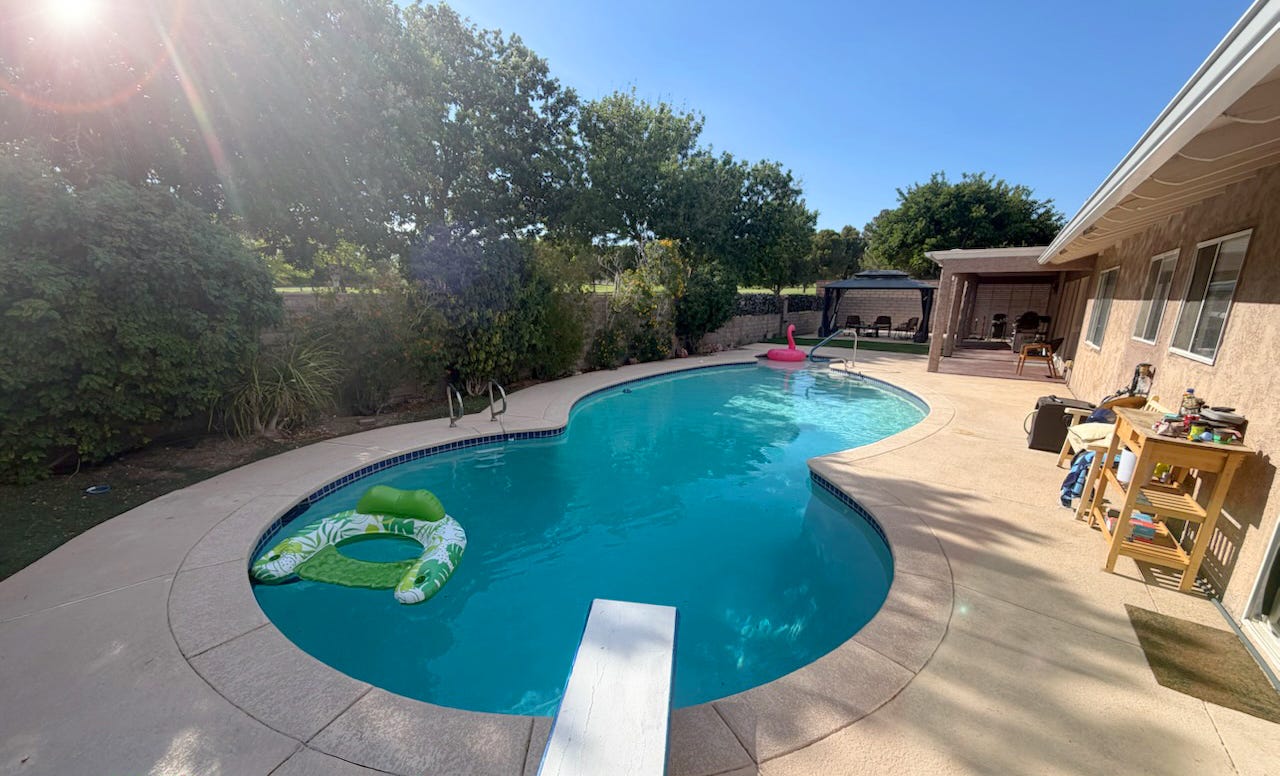A sunlit backyard with a kidney-shaped swimming pool, a diving board, pool floats, a shaded gazebo, and patio seating, ringed by trees and a block wall under a clear blue sky. A sunlit backyard with a kidney-shaped swimming pool, a diving board, pool floats, a shaded gazebo, and patio seating, ringed by trees and a block wall under a clear blue sky.