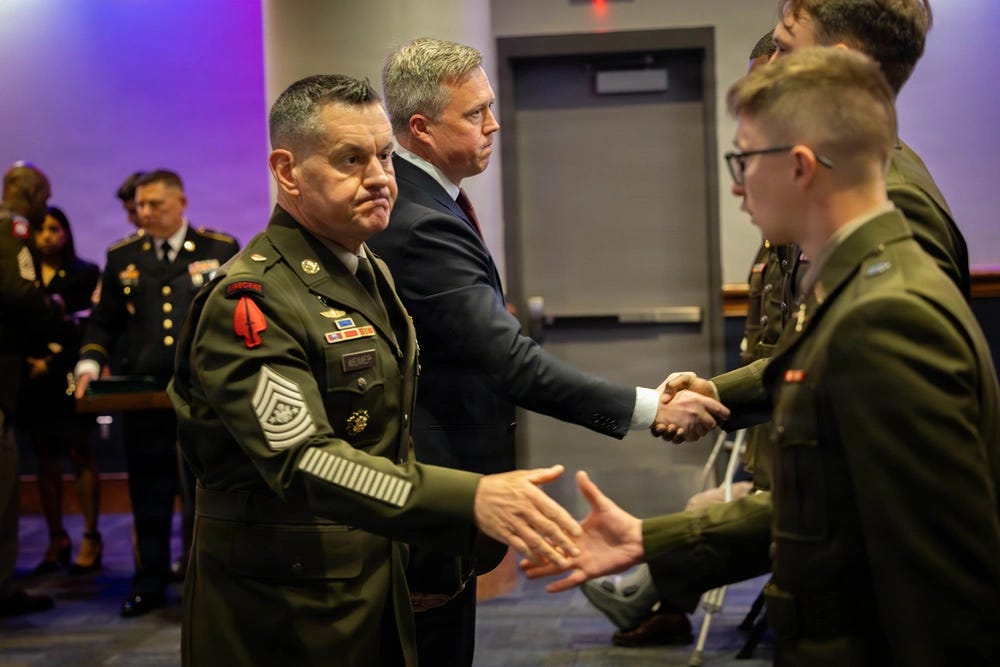 An indoor ceremony scene where a senior U.S. Army service member in dress uniform reaches out to shake hands with a young soldier in uniform, while a man in a suit shakes hands with another uniformed participant. Several other military personnel stand in the background under purple lighting, with a closed door and conference-room setting behind them.
