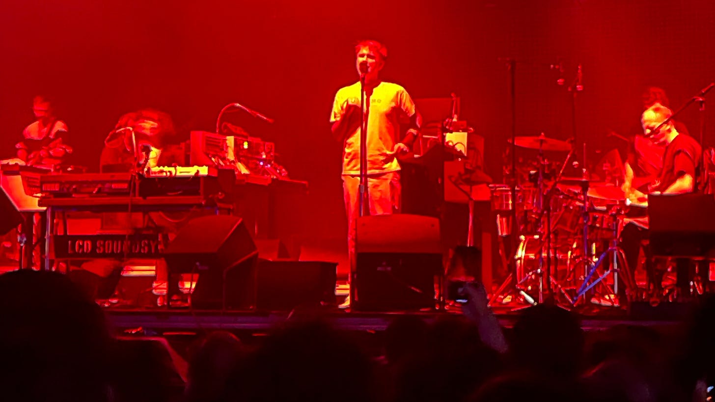 Blurry image bathed in warm light showing LCD Soundsystem performing.