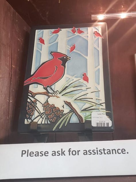 A cardinal mosaic in a shop with "please ask for assistance" under it, a cardinal in a tree sculpture in a park, and a cardinal stencil on a silver car door in a parking lot.