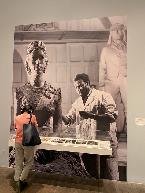 pictures taken at the gallery. Image 1 is of a door with carvings of people. Image 2: a wide picture of the galleries with wooden sculptures. Image 3: An image of a Nigerian sculptor at work in Buckingham Palace