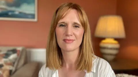 BBC Michelle Guy sitting on a sofa. She has shoulder length strawberry blonde hair, wearing a white linin shirt. A table with a lamp on it is behind her and a picture is hanging on the wall.