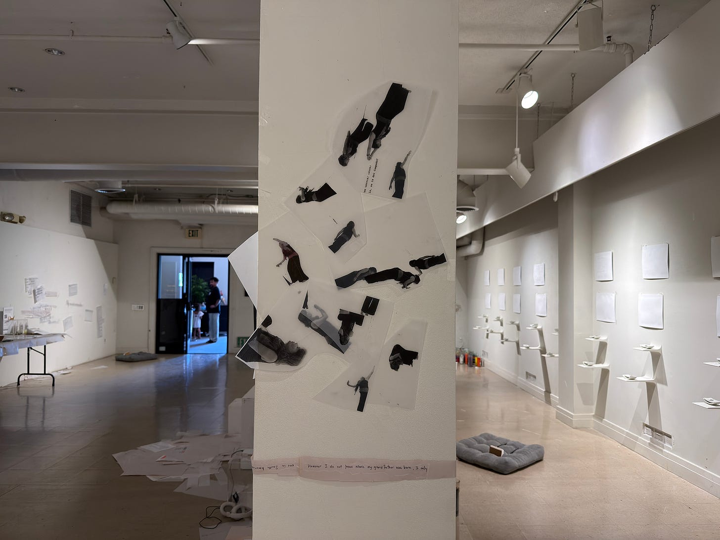 A white structural column hung with irregular sheets of vellum, each carrying cut-out printed figures of a single person in different postures — bending, crouching, reaching, lying down. Most are grayscale; one is in color. The sheets hang from fine monofilament lines and are fixed with strips of clear tape. Past the column, the gallery recedes: pale works on the far wall, a doorway, a rug on the floor.
