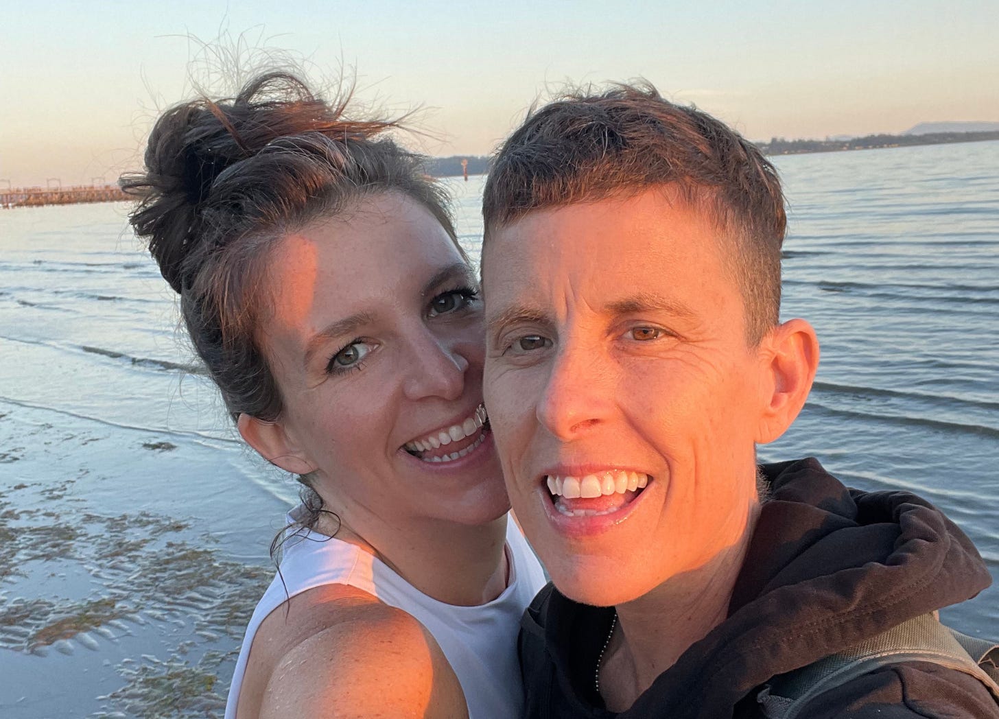 Andrea Gibson and Megan Falley smiling big with the sunshine glowing on their faces standing in front of a sunset filled lake and light blue sky. 
