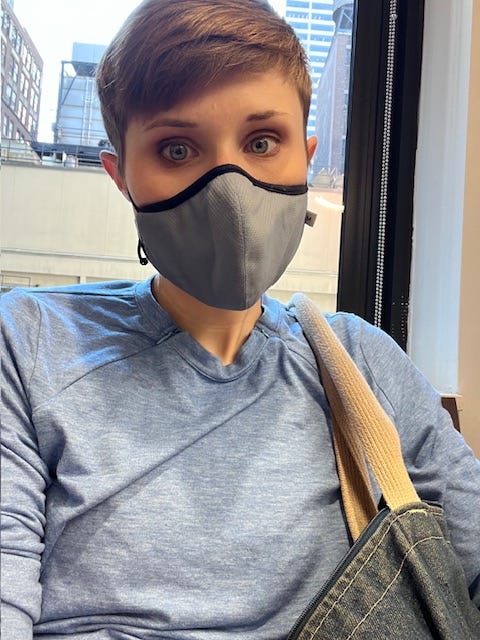 Selfie of the writer (white person, blue shirt, blue medical mask) sitting in front of window in waiting room. 