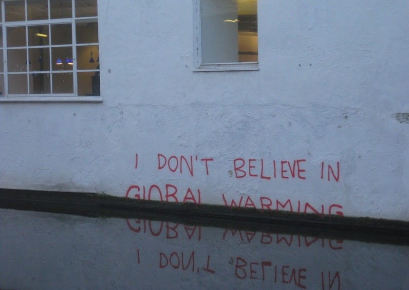 Photo of a white building wall along a still canal, with the phrase 'I don't believe in / global warming' in red spray paint at the level of the water. To simulate flooding, the bottom half of the letters in 'global warming,' are partly cut off, as if just below the surface of the canal. Photo of a white building wall along a still canal, with the phrase 'I don't believe in / global warming' in red spray paint at the level of the water. To simulate flooding, the bottom half of the letters in 'global warming,' are partly cut off, as if just below the surface of the canal.