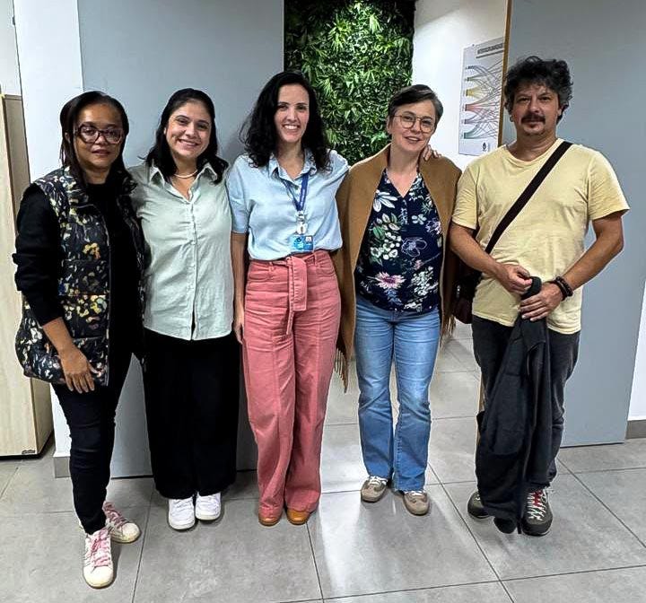 Cinco pessoas em pé lado a lado em um ambiente interno com piso cinzento, paredes brancas e uma parede verde com plantas, sendo da esquerda para a direita: pessoa com cardigan preto com padrão floral dourado e calça preta, pessoa com camisa branca clara e calça preta, pessoa com camisa azul claro, calça rosa e crachá de identificação, pessoa com óculos, blusa com padrão floral em tons escuros e calça jeans, e pessoa com camiseta amarela clara e calça preta com mochila preta atravessada no peito.