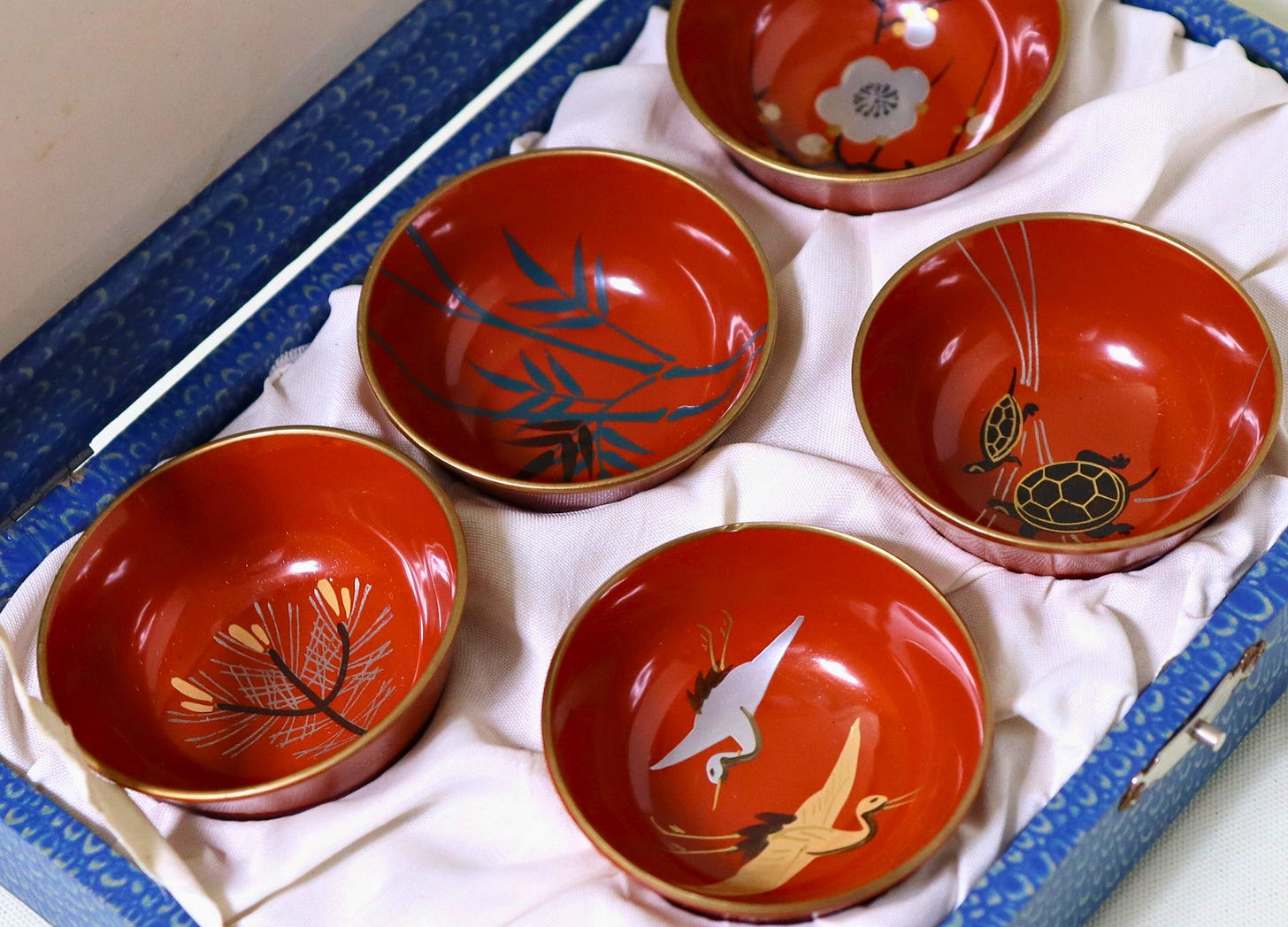 A set of five traditional Japanese lacquered sake cups, each adorned with auspicious motifs like cranes, turtles, pine, bamboo, and plum blossoms, presented in a decorative box.