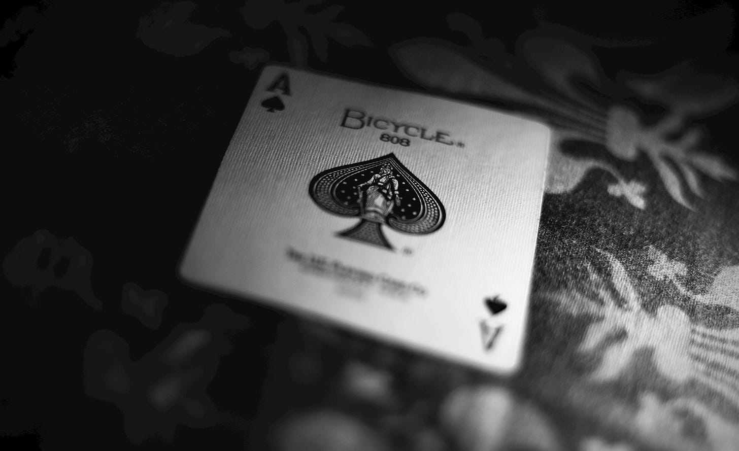 This black and white image displays a close-up, top-down perspective of the bicycle ace of spades from a deck of cards. Located centered on the ace, the central spade pip contains the bust of an intricate statue holding a staff. The top of the card contains the words “BICYCLE” and below it “808” in black capital letters. Small symbols denoting the rank (A) and suit of the card (♠) are found on the top left and bottom right corners. While the card is the main focal point, it is positioned against a background of black and white floral table cloth.