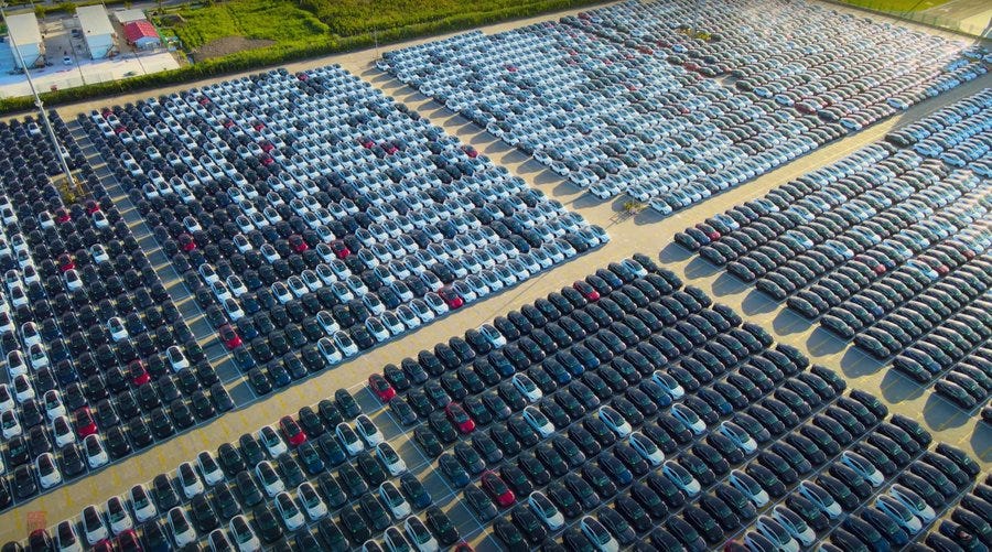 Aerial view of a large outdoor parking lot filled with numerous Tesla vehicles in white, black, and red colors, arranged in organized rows on a paved surface surrounded by green fields and a few buildings in the background under clear daylight.