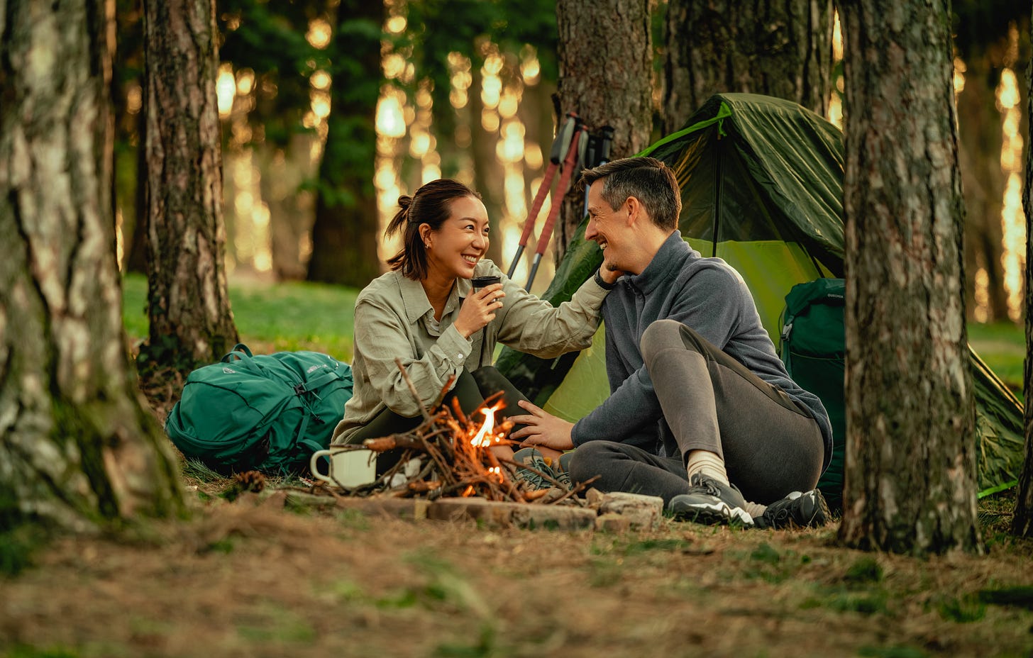 Couple camping in the woods enjoying a glass of wine by the fire.