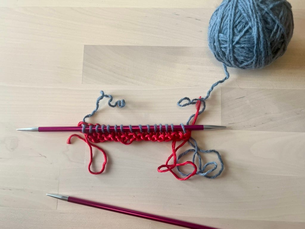 : A knitting needle with rows of red waste yarn below and one row of blue-grey main yarn being worked, on a wooden surface with a ball of blue-grey yarn above.