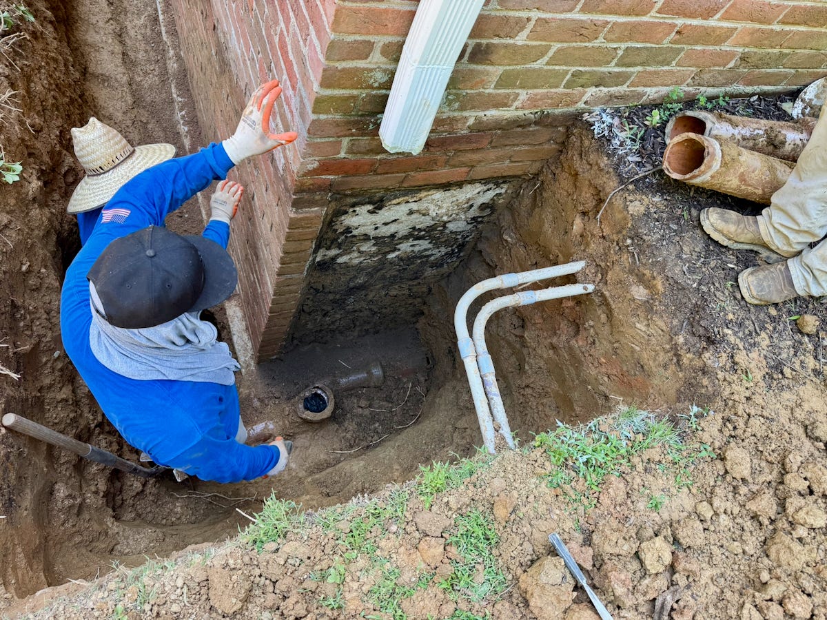 Workers stand in a deep trench beside a brick house, exposing the foundation and pipes for basement waterproofing.
