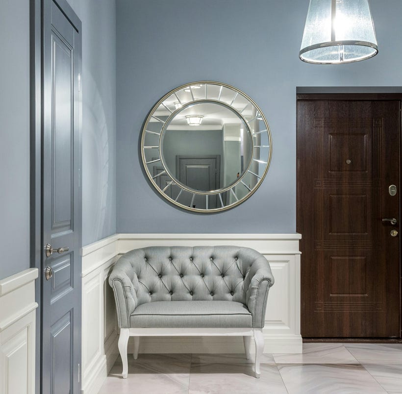 A square image of a hallway with two doors: a deep brown wood one on the right, and a medium-blue one on the left wall. The floor is marble, the walls are wedgewood blue on top and white below a chair rail and fancy wainscoting. In the center of the image are a round gilt mirror on the wall and below it, a tufted blue bench/chair in a place where no one would ever reasonably sit. It looks like your rich grandmother's house in the '80s, except it's all new and therefore somehow creepier.