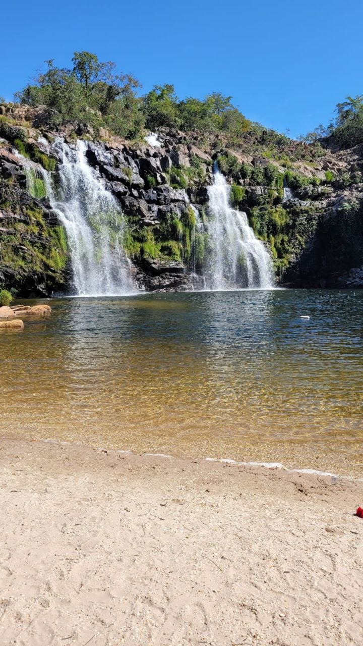 Uma cachoeira baixa mas larga ao fundo, uma área extensa para banho e uma pequena faixa de areia em primeiro plano.