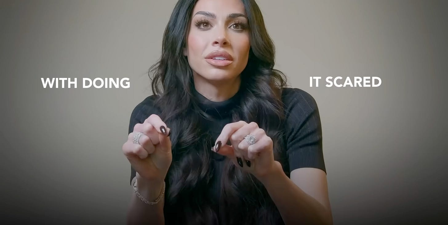 Leila Hormozi sits facing the camera, mid-sentence, with both hands raised in a subtle gesture. She wears a black top and has long, wavy hair. On-screen text appears on either side of her: “WITH DOING” on the left and “IT SCARED” on the right — highlighting the key phrase from her quote about confidence.