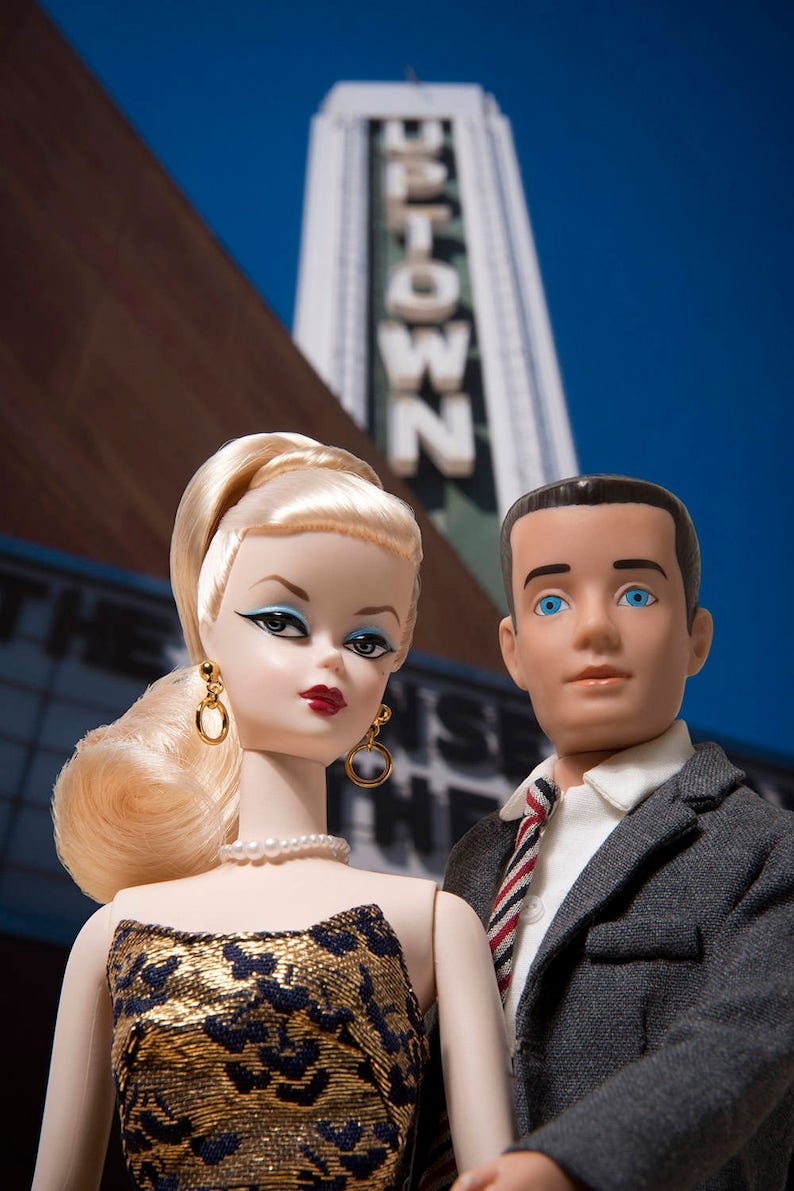 May include: A blonde doll wearing a gold and black dress and a pearl necklace. A doll with brown hair wearing a gray suit and a red and white striped tie. The dolls are standing in front of a sign that says 'UPTOWN'.
