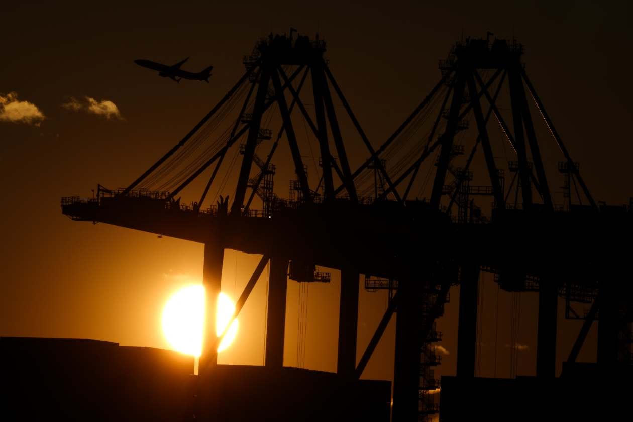Silhouette of cranes at Port Newark Container Terminal with a sunset in the background and a plane flying overhead.