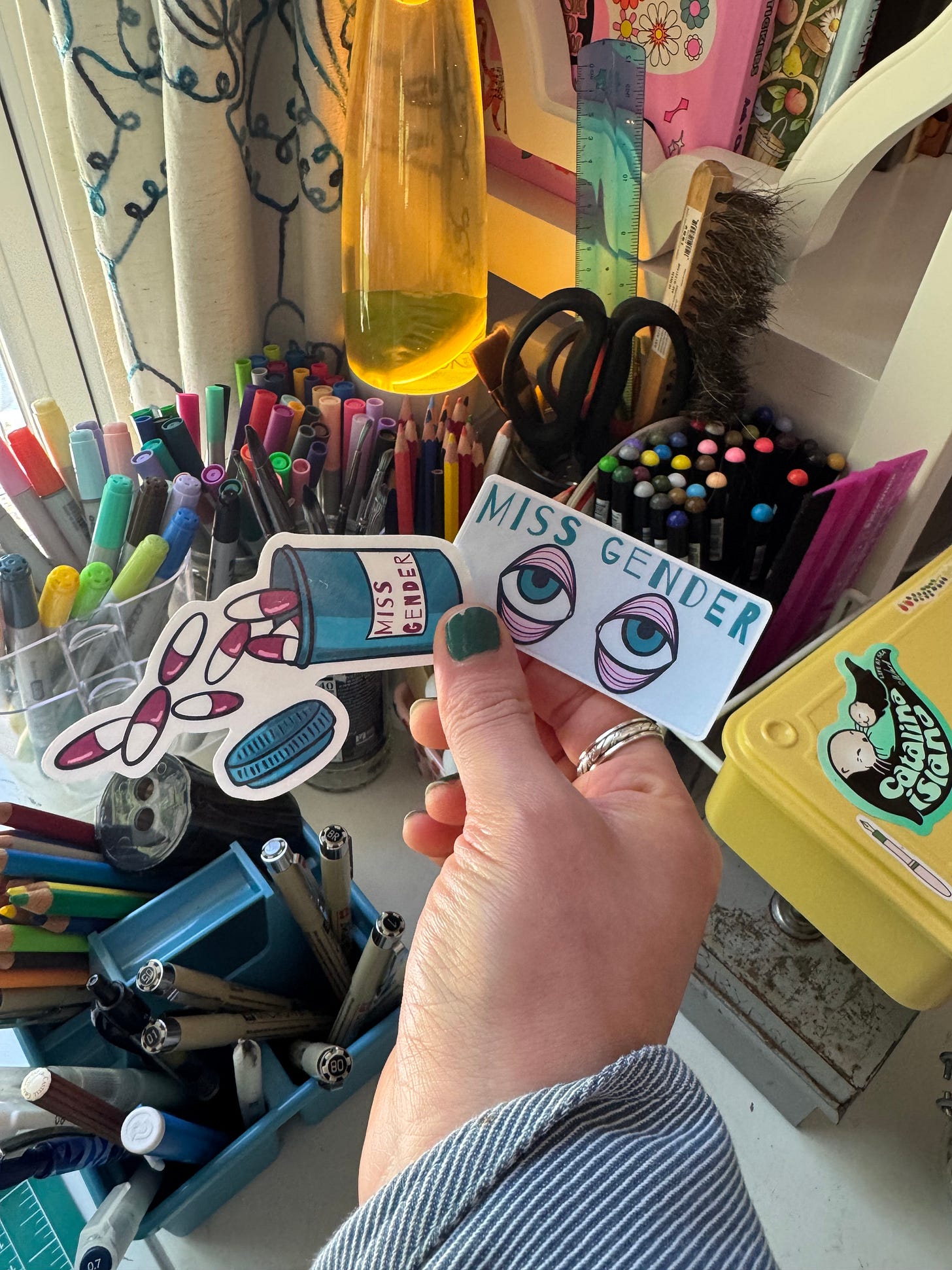 My hand holding two stickers that both read "Miss Gender." One is an illustration of a spilling pill bottle and the other is an illustration of large eyes drawn in my style.