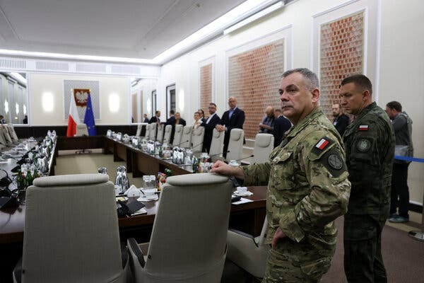 A man in military fatigues rests his right hand on the back of a chair, one of many set up around a conference table. Others in suits and uniforms also stand around the table.