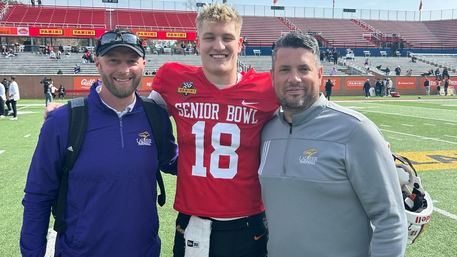 Three people, one in the middle in a red, black and white Senior Bowl football uniform, pose and smile for a photo on a football field. Three people, one in the middle in a red, black and white Senior Bowl football uniform, pose and smile for a photo on a football field.