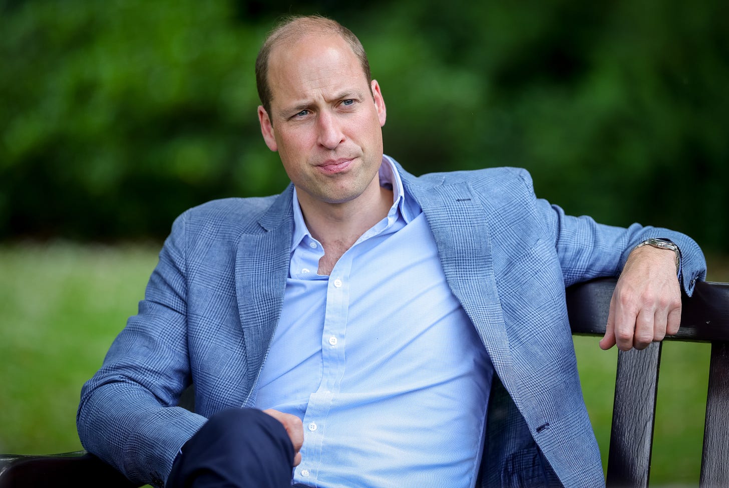 Prince William setting on a bench wearing a blue shirt and jacket