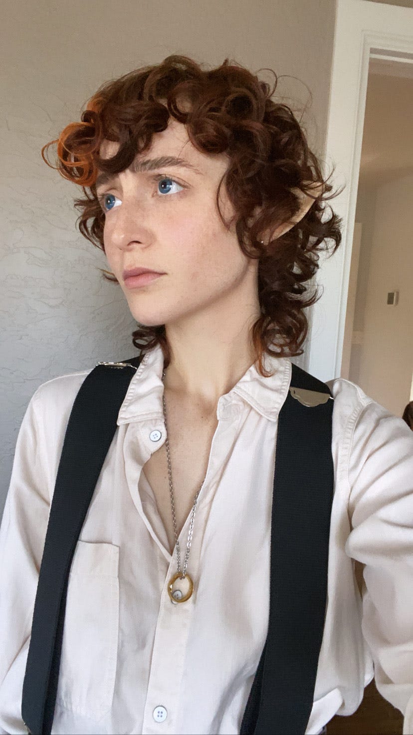 Jo O'Neill as Frodo baggins for halloween, wearing blue contacts, a cream button down shirt, replica of the One Ring, and black suspenders. they are making the concerned Frodo face.