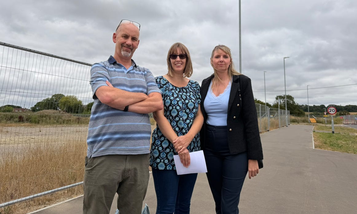    Three adults stand on a paved walkway beside a fenced-off area under an overcast sky. One person has their arms folded, another holds a sheet of paper, and the third stands slightly behind them. Behind the group is Heras fencing enclosing dry grassland, with a 20 mph road sign visible on the adjacent road.