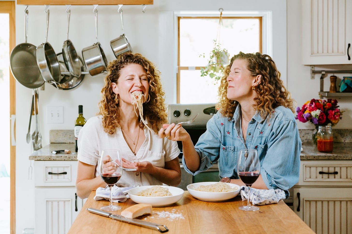 two sisters sitting in a kitchen smiling and enjoying a plate of spaghetti