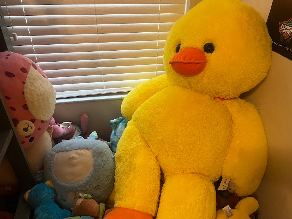 Photo of a giant stuffed duck resting on top of a pile of smaller stuffed animals in the corner of a bedroom.