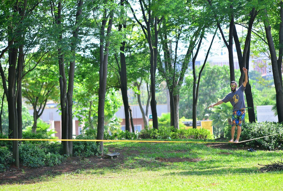 Pessoa praticando slackline em um parque arborizado, equilibrando-se sobre uma fita suspensa entre árvores, com vegetação ao redor e edificações ao fundo.