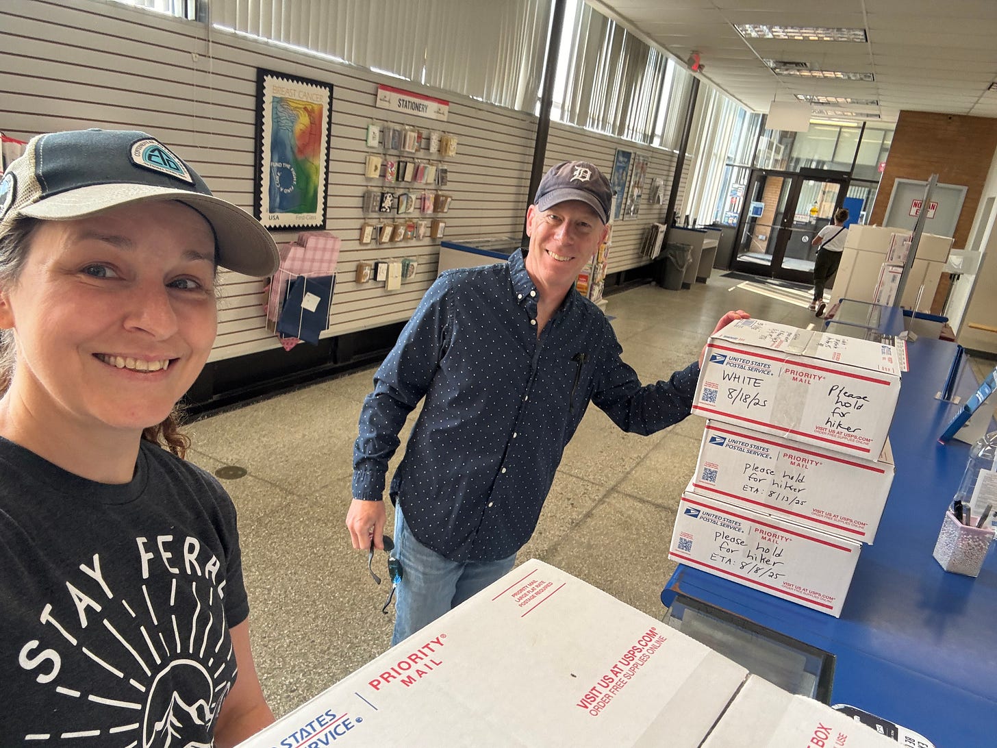 The author and her husband are standing in the post office with their resupply boxes for their upcoming hike, smiling from ear to ear. Almost time to go! 