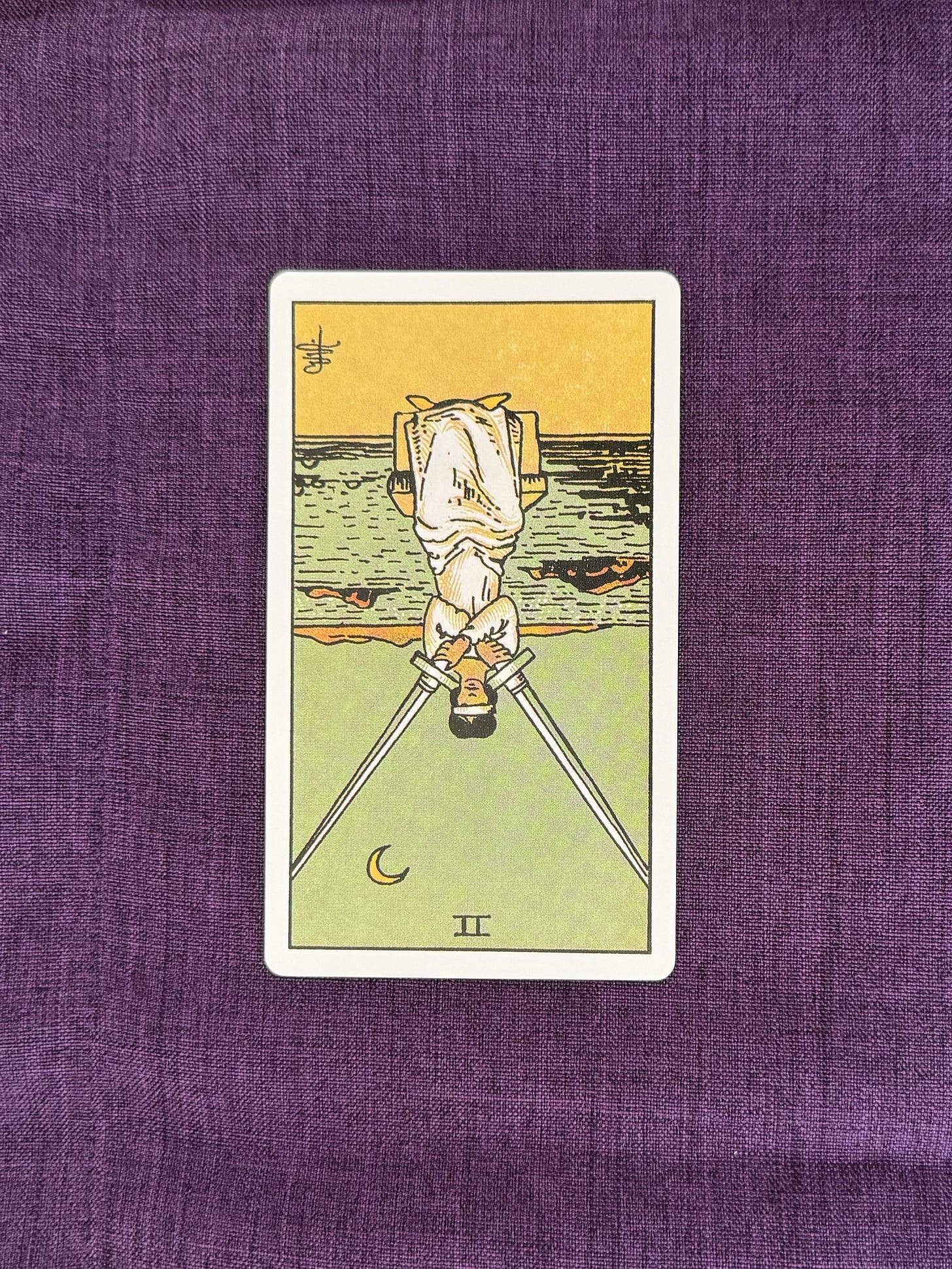Tarot Card is on a purple linen cloth, depicting: A female figure sits blindfolded, as she balances two swords across her shoulders. Behind her is a large body of water and above her is the moon.