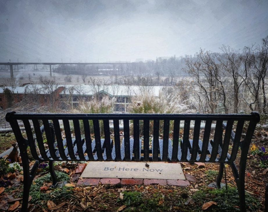Black metal bench seen from behind, looking over a misty James River in Virginia; a stone tablet at the foot of the bench reads "Be Here Now" Black metal bench seen from behind, looking over a misty James River in Virginia; a stone tablet at the foot of the bench reads "Be Here Now"