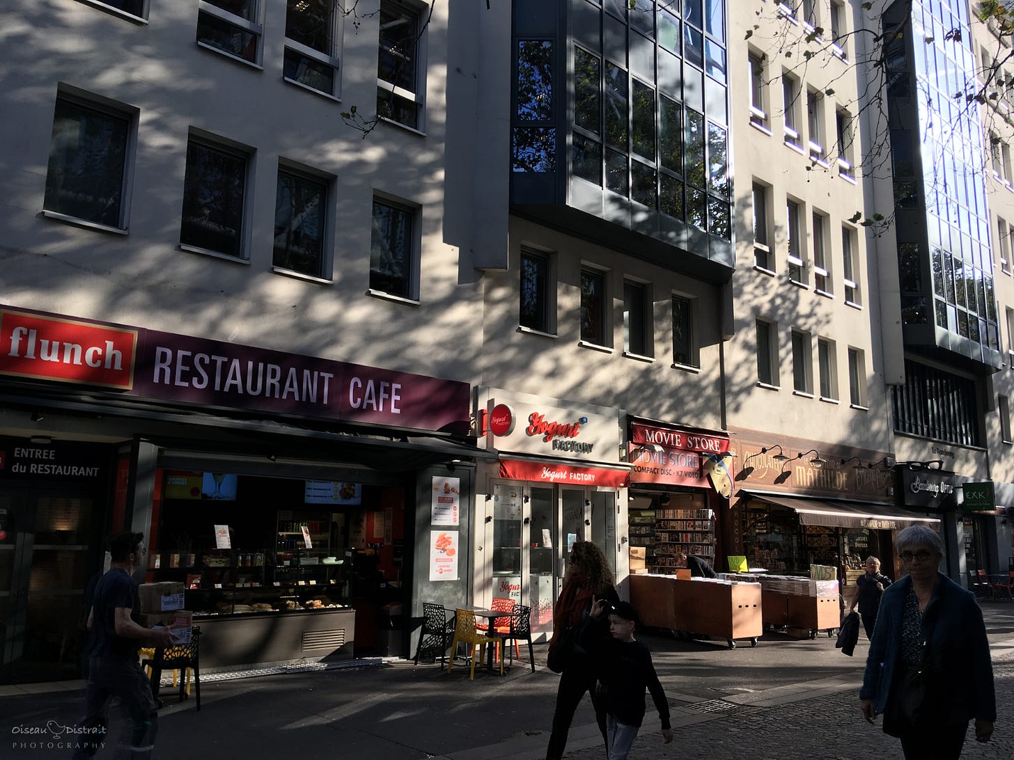 A street view of Paris under a sunny day, pedestrians outside shops & cafe. Tree shadow plays with the sun cast on the building façade. A street view of Paris under a sunny day, pedestrians outside shops & cafe. Tree shadow plays with the sun cast on the building façade.