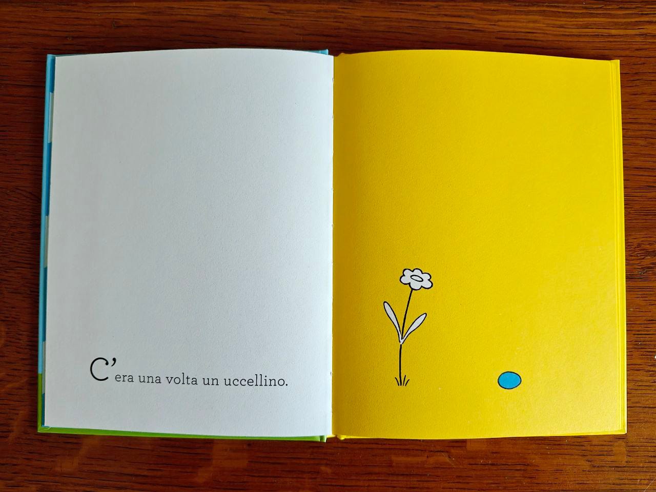 Foto di una doppia pagina del libro "L'uovo felice" appoggiata su un tavolo di legno marrone. A sinistra pagina bianca e le parole "C'era una volta un uccellino.". A destra pagina gialla con un fiore bianco e un uovo azzurro. Foto di una doppia pagina del libro "L'uovo felice" appoggiata su un tavolo di legno marrone. A sinistra pagina bianca e le parole "C'era una volta un uccellino.". A destra pagina gialla con un fiore bianco e un uovo azzurro.