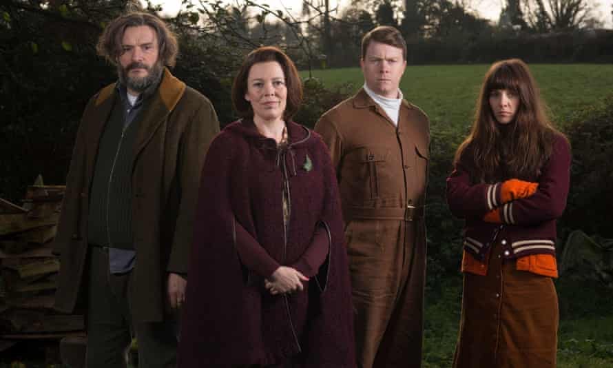 A dark picture of four people facing the camera. L-R is a middle-aged man with floppy hair (Julian Barrett), a middle-aged woman with brown hair and a burgundy cloak (Olivia Coleman), a younger man in a brown jumpsuit (Daniel Rigby), and a younger woman with a heavy dark fringe and her arms crossed (Sophia Di Martino), and in the background a stretch of green grass and wall of stones