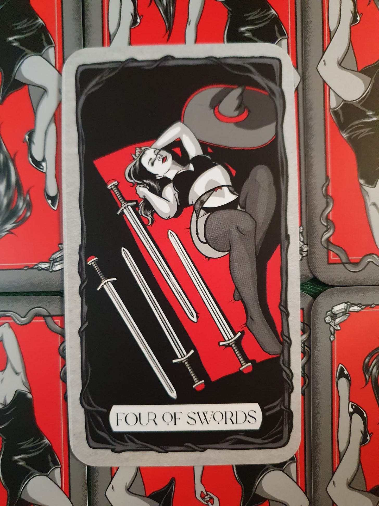 The Four of Swords tarot card featuring a reclining woman dressed in black lingerie and stockings, surrounded by four silver swords. A dark, horned shape looms above her. The artwork features a striking red, black and grey gothic aesthetic. The Four of Swords tarot card featuring a reclining woman dressed in black lingerie and stockings, surrounded by four silver swords. A dark, horned shape looms above her. The artwork features a striking red, black and grey gothic aesthetic.
