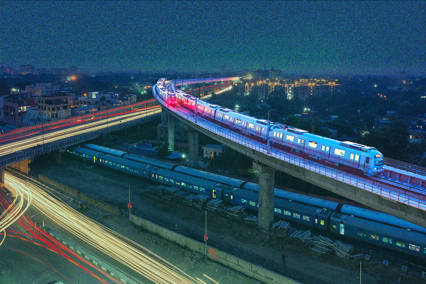 at night, an elevated train moves above a ground train with the lights of cars on roads around them