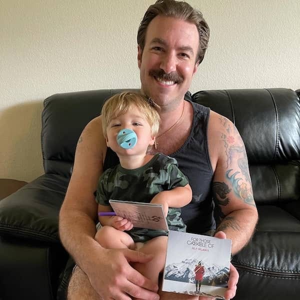 A man holds a young child on his lap, both smiling. The man has a mustache and is wearing a sleeveless shirt, while the child is dressed in a camouflage outfit and has a pacifier. They are sitting on a black leather couch, and the man is displaying an album cover titled 'FOR THOSE CAPABLE OF.' A man holds a young child on his lap, both smiling. The man has a mustache and is wearing a sleeveless shirt, while the child is dressed in a camouflage outfit and has a pacifier. They are sitting on a black leather couch, and the man is displaying an album cover titled 'FOR THOSE CAPABLE OF.'