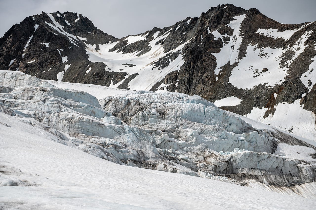 A tiered wall of glacier ice, white and pale blue, banded with dark debris lines, stretches across the frame below a dark rock peak streaked with snow gullies. Smooth snowfields lap against the base of the ice cliffs in the foreground, under a flat grey sky.