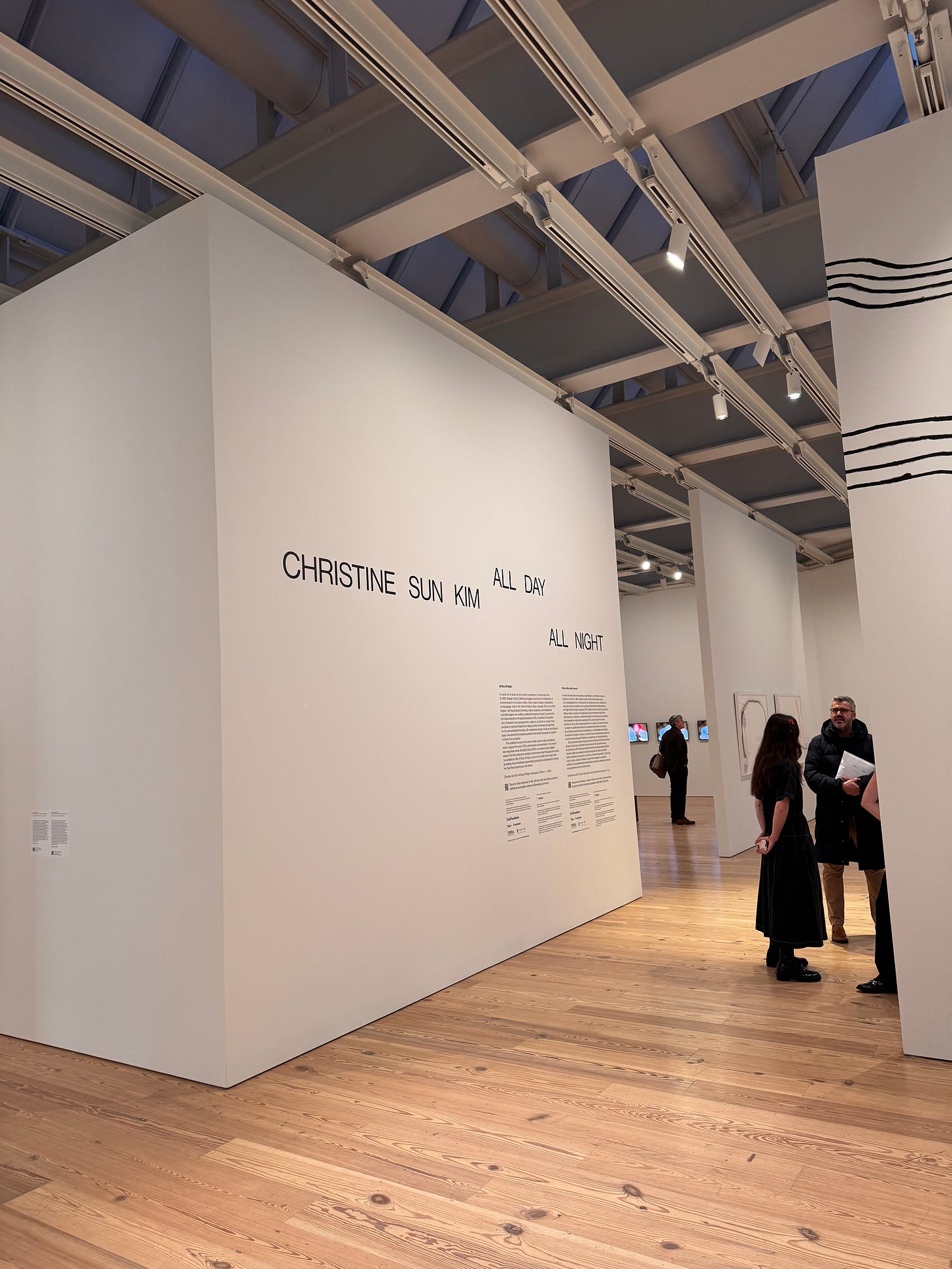 "Christine Sun Kim: All Day All Night" in all caps across a white wall at the Whitney Museum of American Art. Exhibition halls in the background, some people gather on smooth wooden floors chatting.