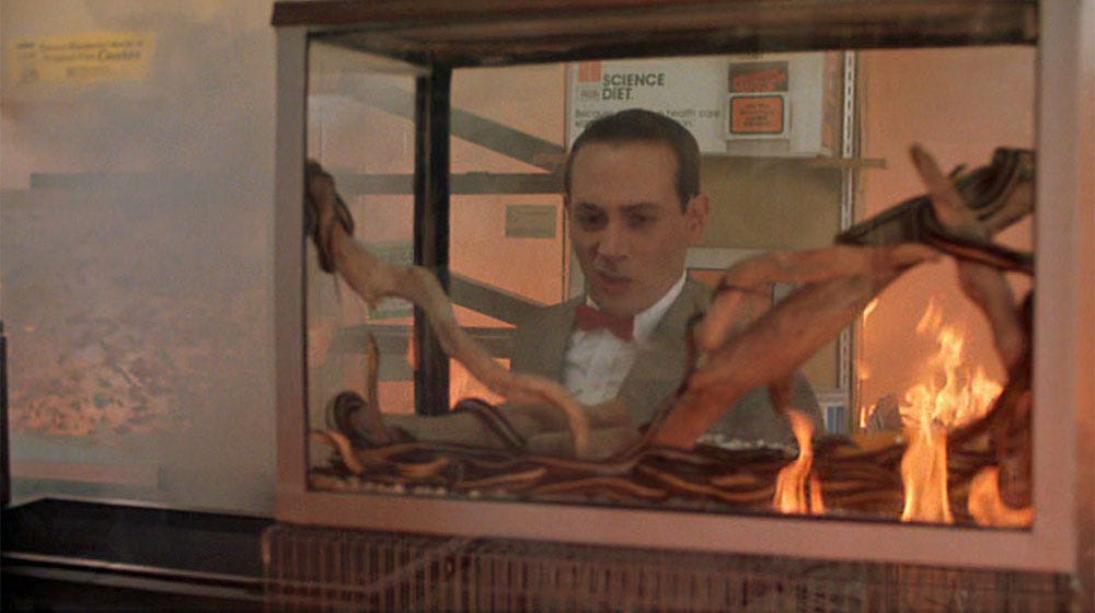 A still from Pee Wee's Big Adventure (1985) where Pee Wee is looking at a terrarium of snakes in disgust while a fire rages in the pet shop.