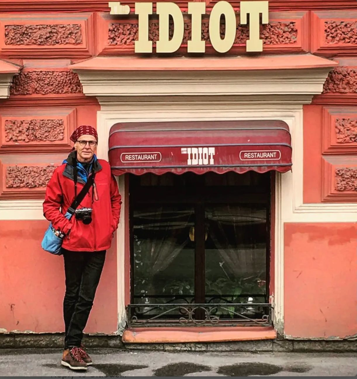 A person in a red jacket stands in front of a coral-pink building with "The Idiot" restaurant sign above the entrance on a grey winter street in St. Petersburg.