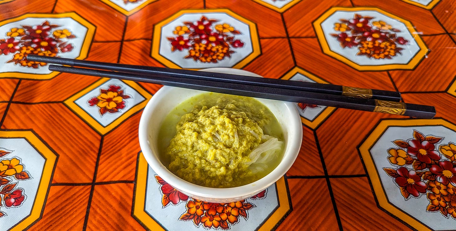 A photo of rice noodle soup sitting on an orange-colored table, black chopsticks resting across the white bow. 