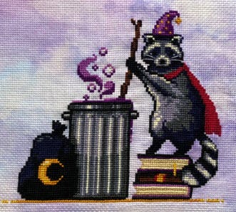 A cross stitch of a raccoon wearing a cape and starry wizard hat, stirring a trashcan with a long stick.  Magical bits are wafting from the trashcan and a black garbage bag with a crescent moon on it lies nearby.