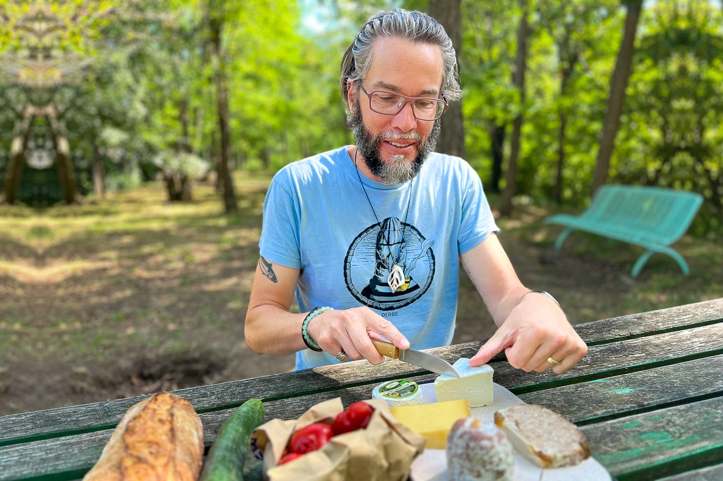 Patrick Hemminger mit Brille und Bart sitzt an einem verwitterten Gartentisch im Grünen und schneidet ein Stück Käse. Vor ihm liegen Brot, Gurke, Tomaten, weiterer Käse und aufgeschnittene Brotscheiben; im Hintergrund stehen Bäume und eine türkisfarbene Bank.