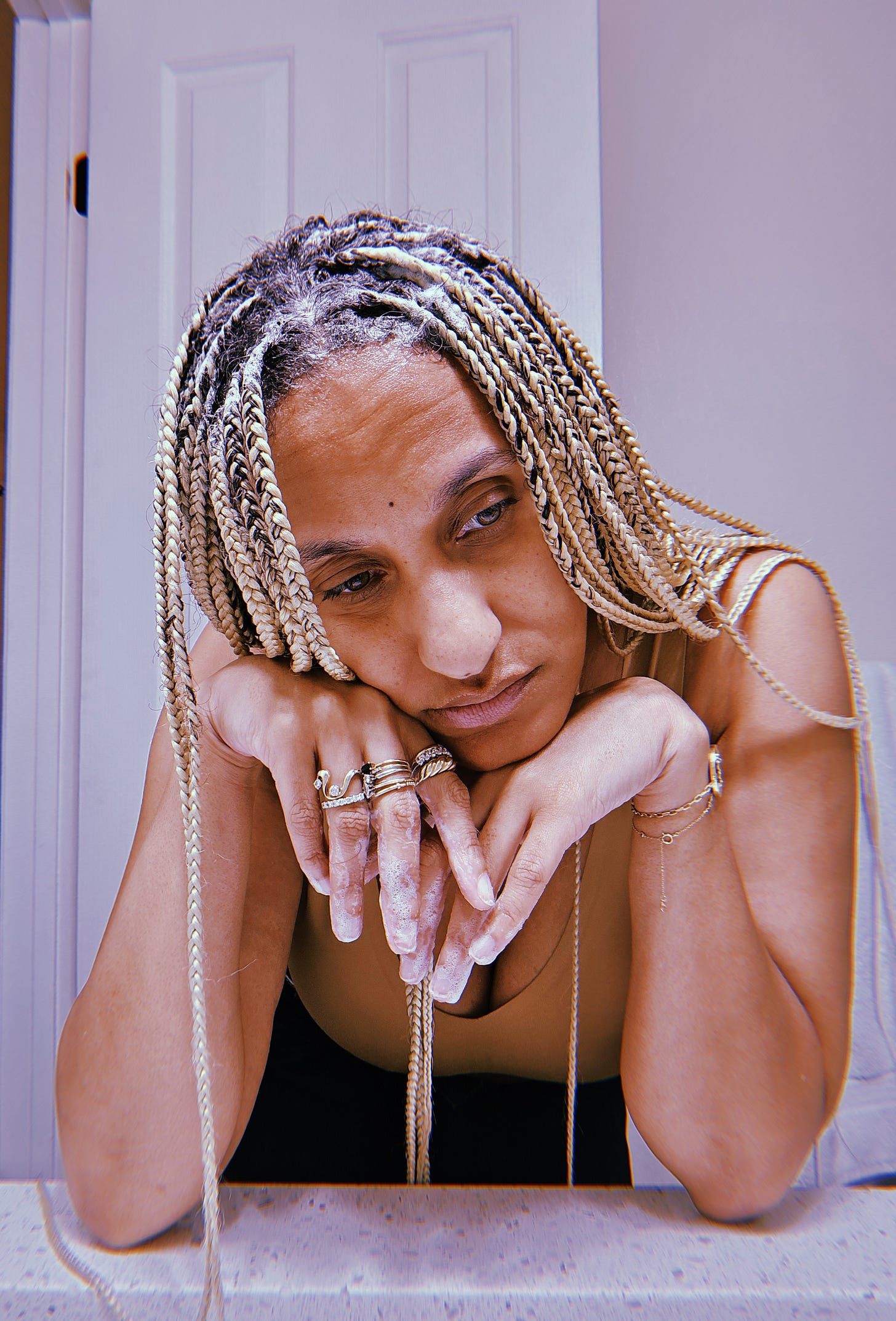 Dr. Cadet is that person who looks stunning when exhausted and feeling like a failure and yes it’s not fair. She rests her elbows on a white bathroom countertop with her fingertips are covered in bubbles from shampoo as she takes a break from washing her golden braids cuz disability. Her head is propped up by her hands with gold  rings from her accomplishments looking to one side. Pops of her hazel eyes look to the distance as she wears a nude bra giving a peek of her award winning cleavage.