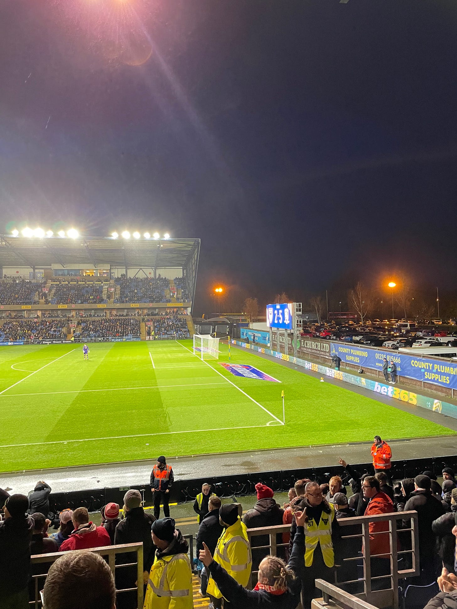 The match in progress, the open end of Oxford's stadium with the scoreboard reading '2-5'