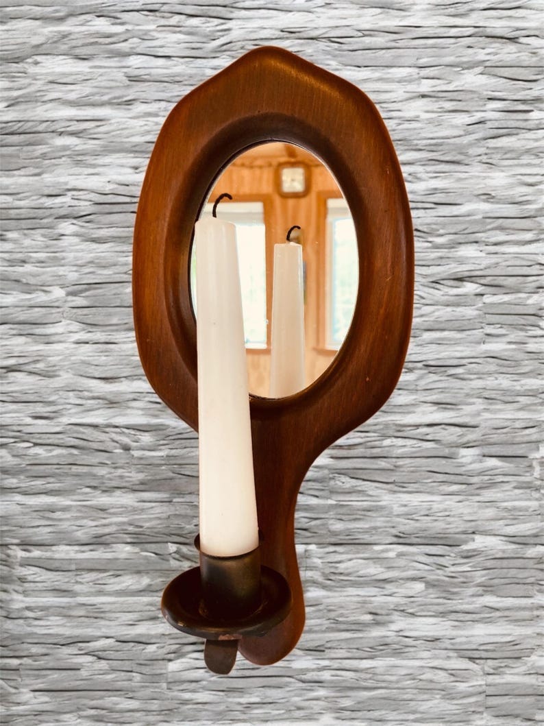 May include: A wooden wall-mounted mirror with a candle holder. The mirror is oval-shaped and has a dark wood frame. The candle holder is on the bottom of the mirror and has a brass finish. May include: A wooden wall-mounted mirror with a candle holder. The mirror is oval-shaped and has a dark wood frame. The candle holder is on the bottom of the mirror and has a brass finish.